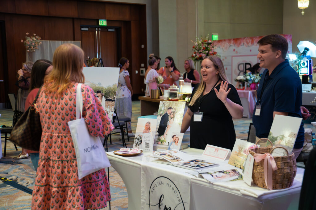 Wedding professionals greet and chat with an attendee at a bridal show booth, displaying albums, framed wedding photos, and printed materials while the guest carries a NOW Weddings tote bag.