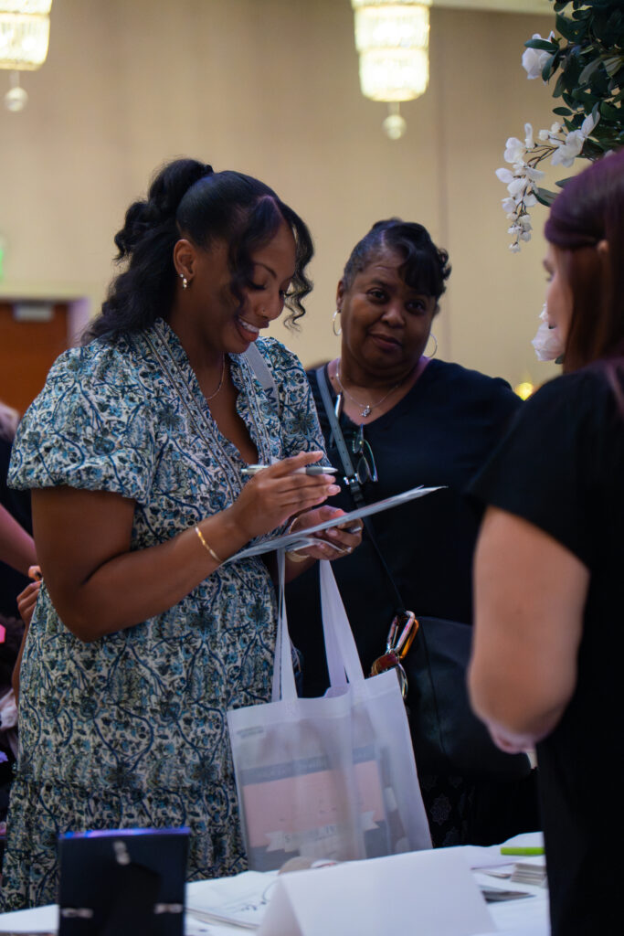 Attendee completing a form at a bridal show booth while holding a NOW Weddings tote bag, engaging with a vendor amid floral décor and wedding planning materials.