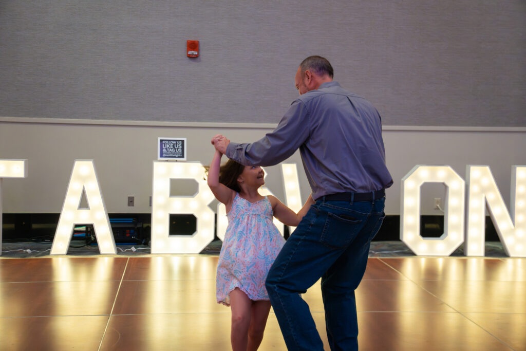 Father and young daughter dancing together on a ballroom floor in front of illuminated marquee letters at a wedding expo.