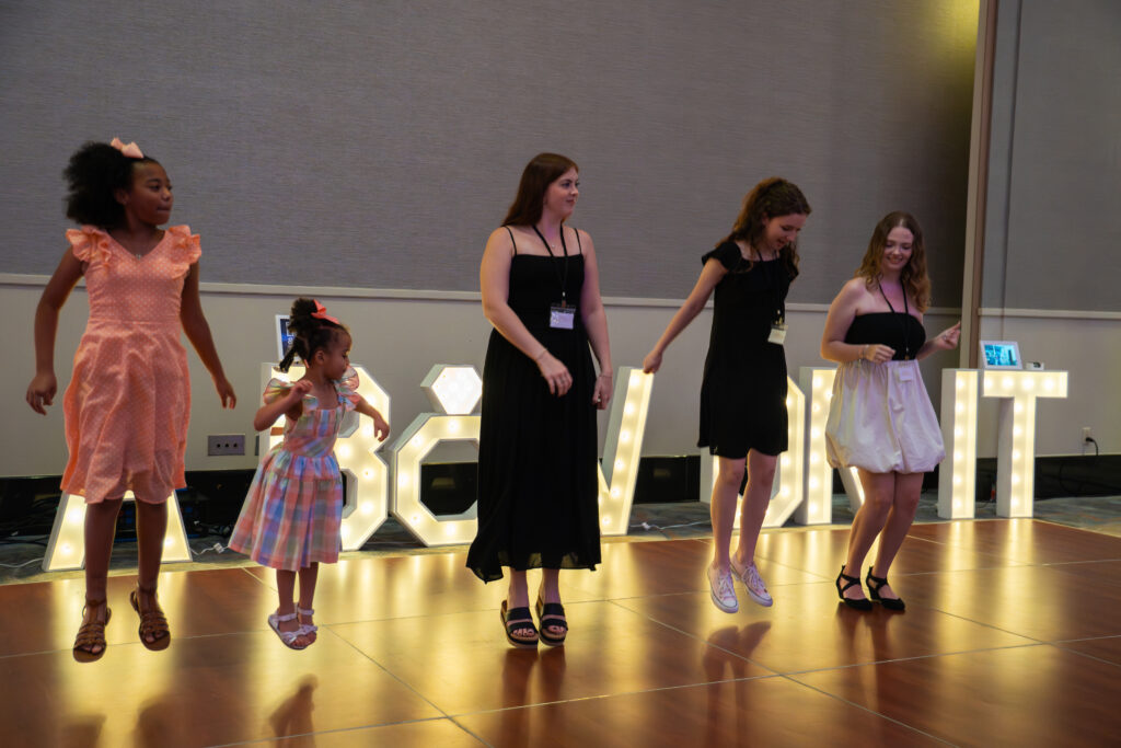 Group of girls and women dance together on a lit dance floor in front of glowing marquee letters at a wedding showcase event.
