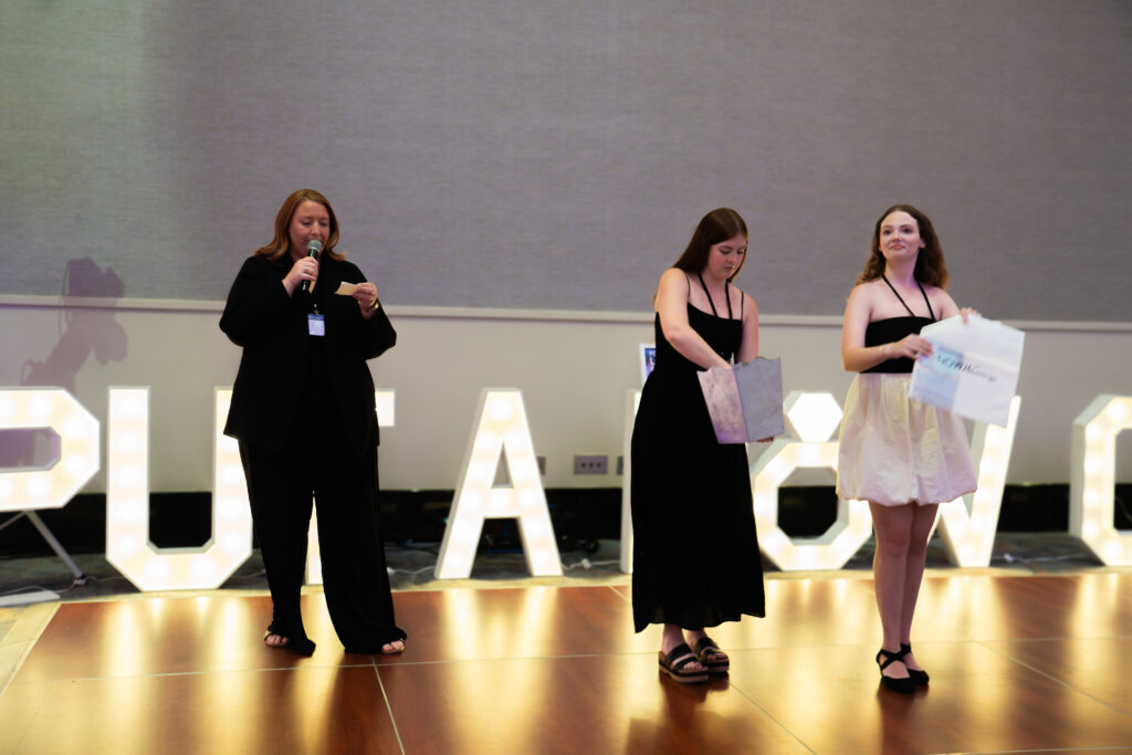 Event host speaking into a microphone on stage while two assistants hold gift bags and signage during a bridal show giveaway.