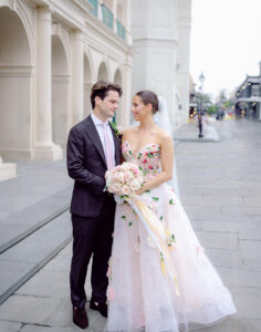 Bride and groom stand together on a New Orleans street, the bride holding a pastel rose bouquet and wearing a floral strapless wedding gown.