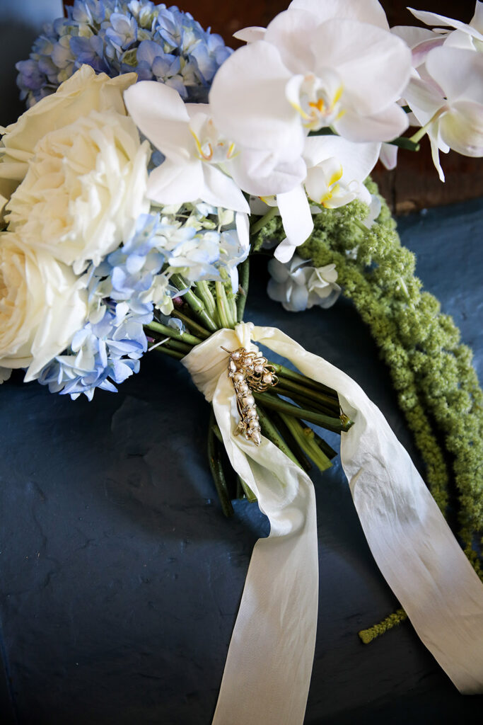 Bridal bouquet featuring white roses, orchids, and soft blue hydrangeas, wrapped with flowing ivory ribbon and a vintage brooch detail.