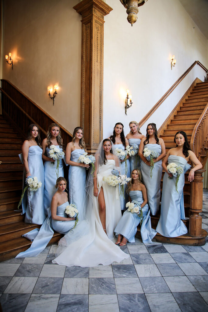 Bride stands on a grand staircase surrounded by bridesmaids in pale blue dresses, each holding white and blue floral bouquets.