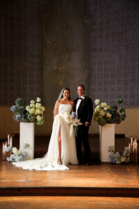 Bride and groom posing together at the altar, featuring modern floral arrangements, candlelight, and a textured wall backdrop in an elegant indoor wedding setting.