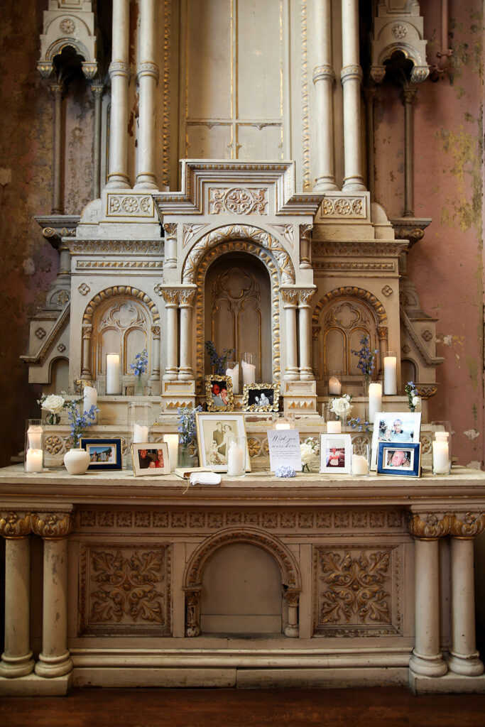 Memorial table with framed family photos, candles, and white and blue florals arranged on an ornate church altar.