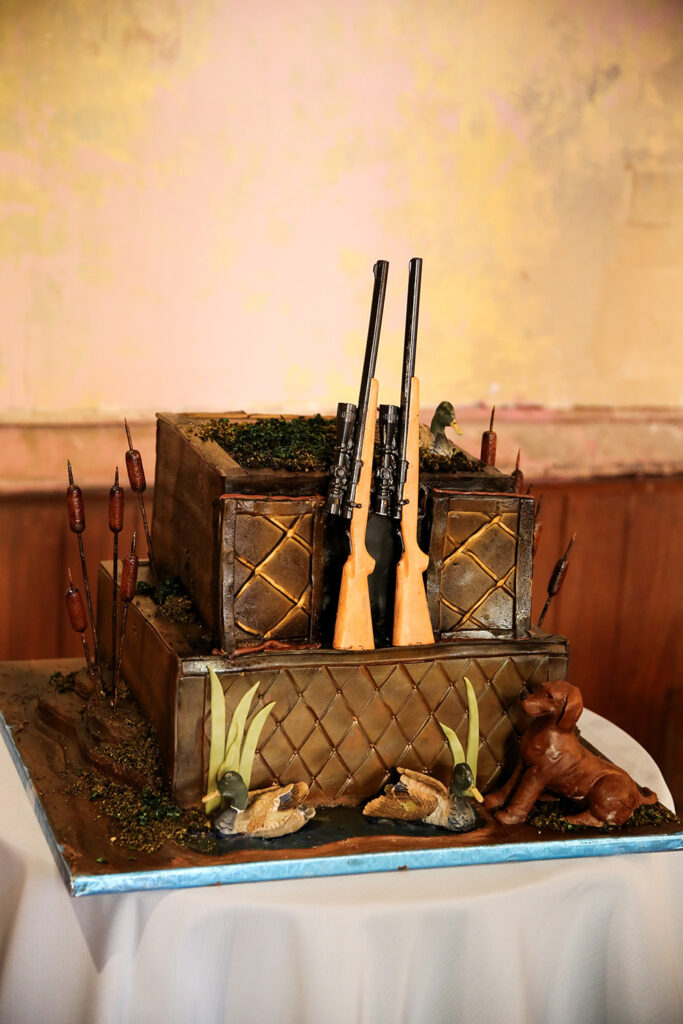Groom’s cake designed with a rustic hunting theme, featuring sculpted rifles, ducks, cattails, and a dog detail.