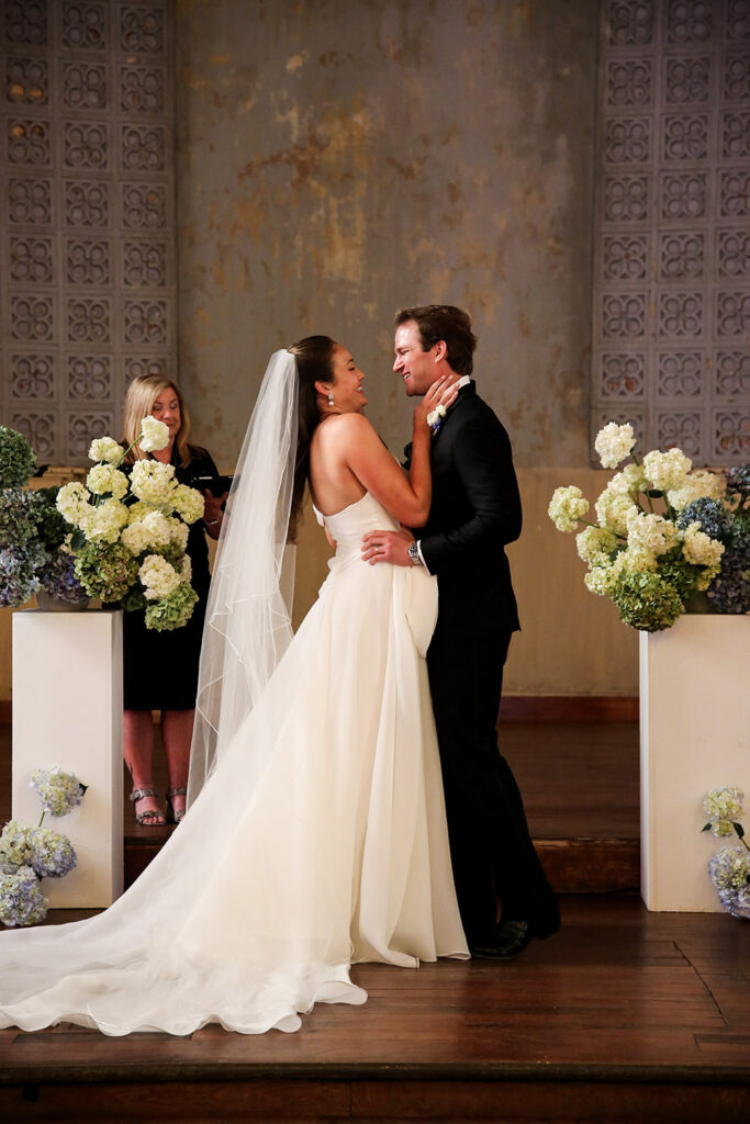 Bride and groom share a joyful moment during their ceremony, surrounded by white and blue floral arrangements at the altar.