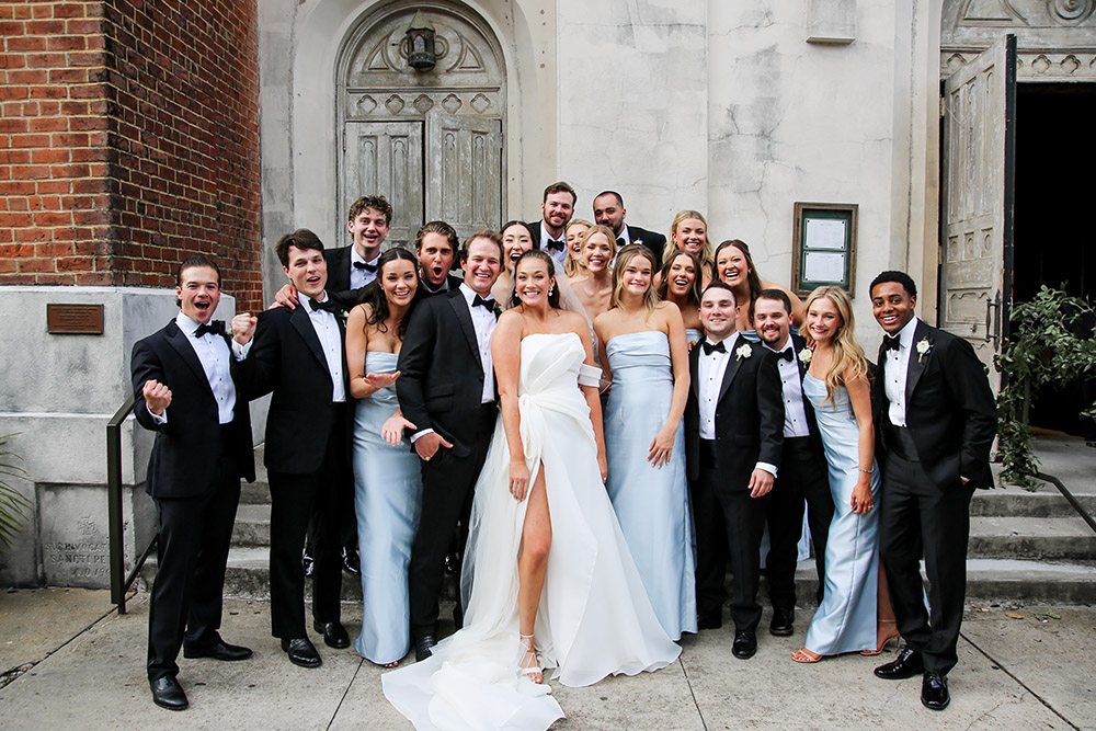 Bride and groom pose with their wedding party outside a church, surrounded by smiling bridesmaids in pale blue dresses and groomsmen in black tuxedos.