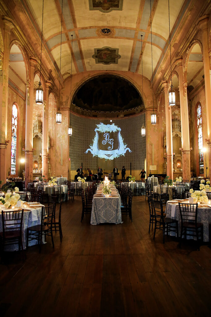 Bride and groom smile while cutting their wedding cake inside a historic venue, surrounded by white and blue floral details.