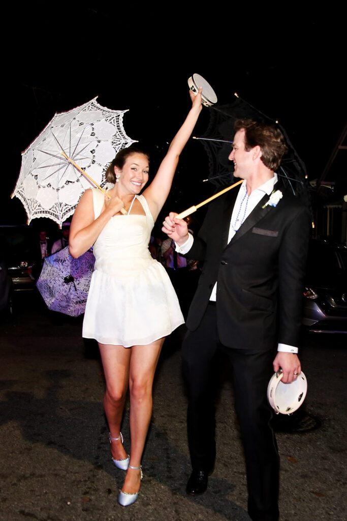 Bride and groom celebrate during a nighttime second line, smiling and dancing with umbrellas and tambourines.