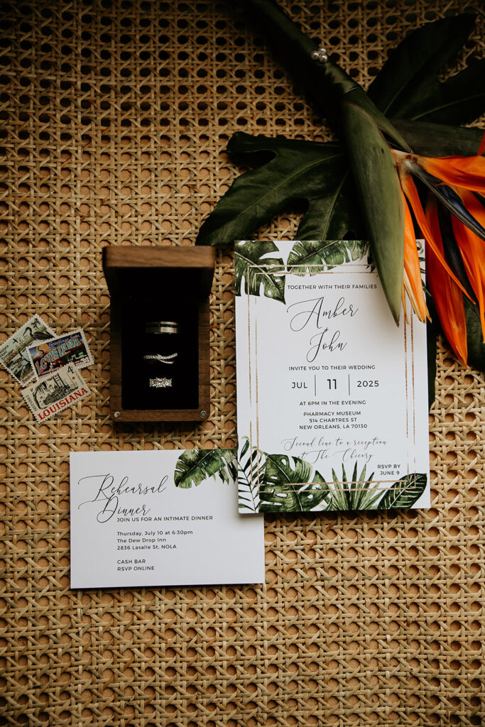 Here’s keyword-rich, ADA-friendly alt text for this image:

**Alt Text:**
Flat lay of tropical-themed wedding details on a woven rattan surface, including a wooden ring box with wedding bands, Louisiana postage stamps, Bird of Paradise flowers, and elegant green-and-white invitations for Amber and John’s New Orleans wedding and rehearsal dinner.
