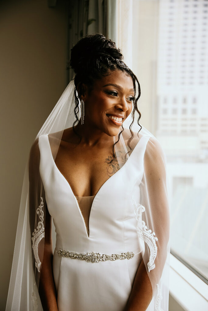 A smiling bride stands by a window in a modern hotel room, wearing a sleek white wedding gown with a deep V-neckline, jeweled belt, and lace-trimmed veil. Her hair is styled in an elegant updo, and she looks out toward the light with soft, natural makeup and sparkling earrings.