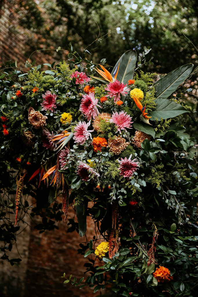 Here is a polished, SEO-friendly alt text option for this floral detail:

**Alt Text:**
Close-up of a vibrant wedding ceremony floral arch featuring tropical greenery, pink and orange flowers, and birds-of-paradise against a brick courtyard backdrop.
