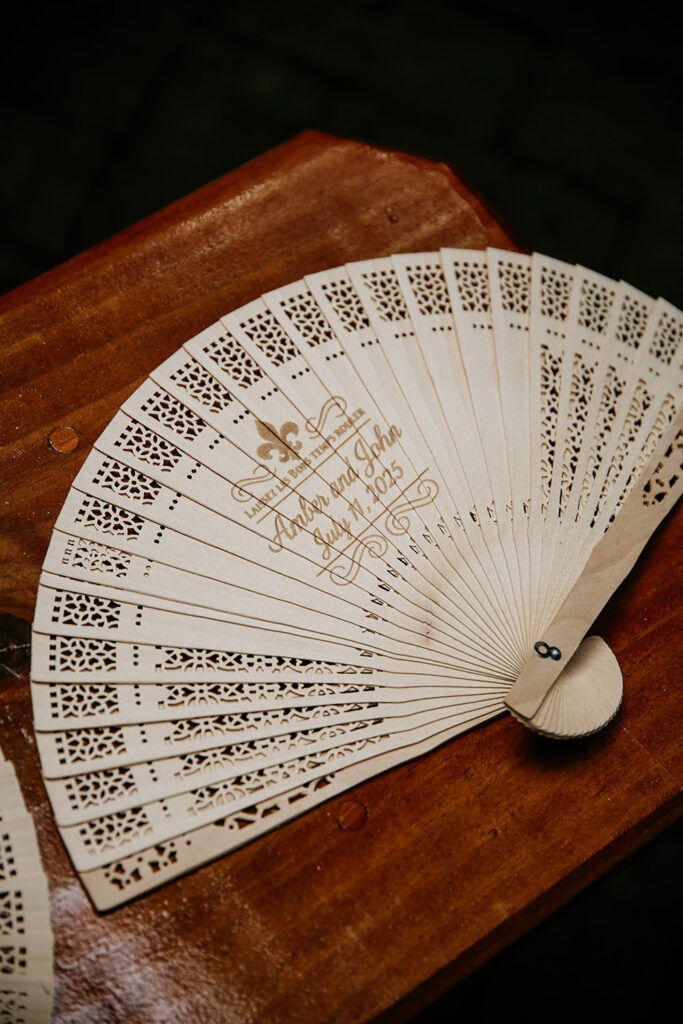Here’s SEO-friendly alt text for this detail shot:

**Alt Text:**
Custom laser-cut wedding fan with Amber and John’s names and July 11, 2025 date displayed on a wooden table as a ceremony favor.
