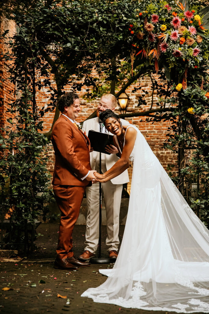 Here’s optimized alt text for accessibility and SEO:

**Alt Text:**
Bride and groom share a joyful laugh during their outdoor wedding ceremony under a lush floral arch at the Pharmacy Museum in New Orleans.
