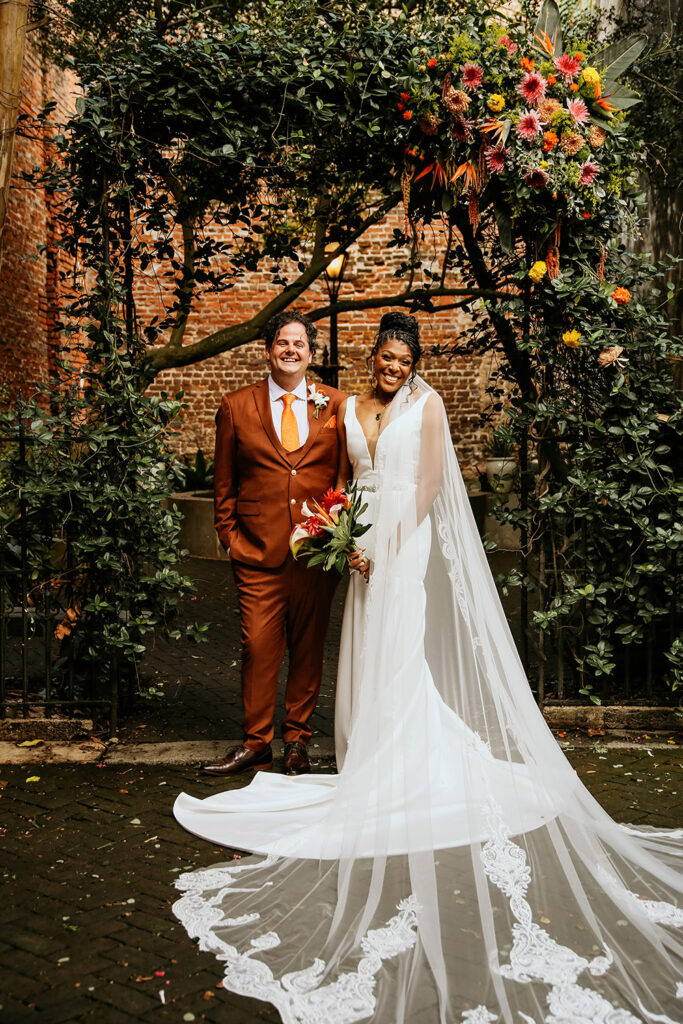 Here’s SEO-optimized alt text for this image:

**Alt Text:**
Bride and groom smiling under a lush floral arch at their New Orleans courtyard wedding, with the bride in a long lace veil and the groom in a rust-colored suit holding a tropical bouquet.

