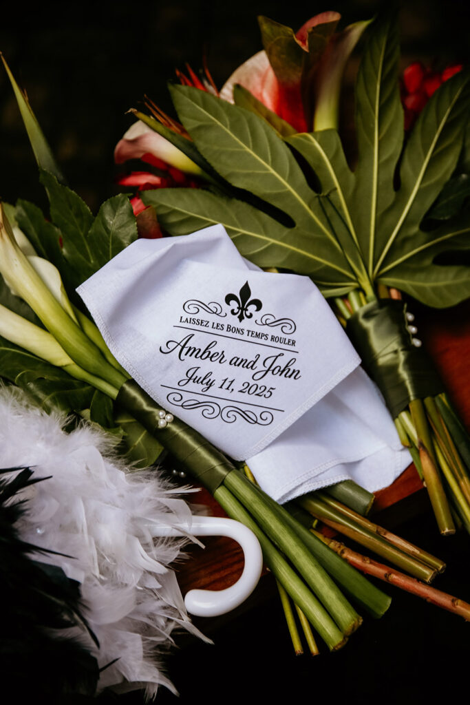 Close-up of wedding florals and a custom white handkerchief printed with “Amber and John, July 11, 2025” and a fleur-de-lis, alongside a white feathered parade umbrella handle.