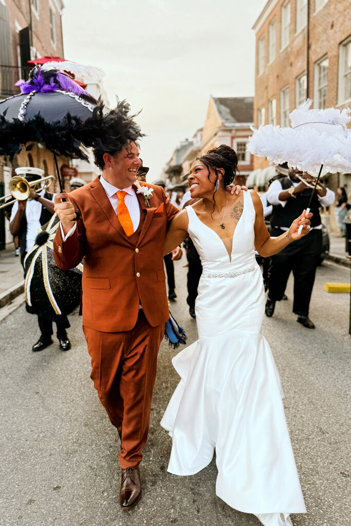 Bride and groom dancing joyfully in a New Orleans second line parade, holding feathered umbrellas and smiling as a brass band celebrates behind them in the French Quarter.