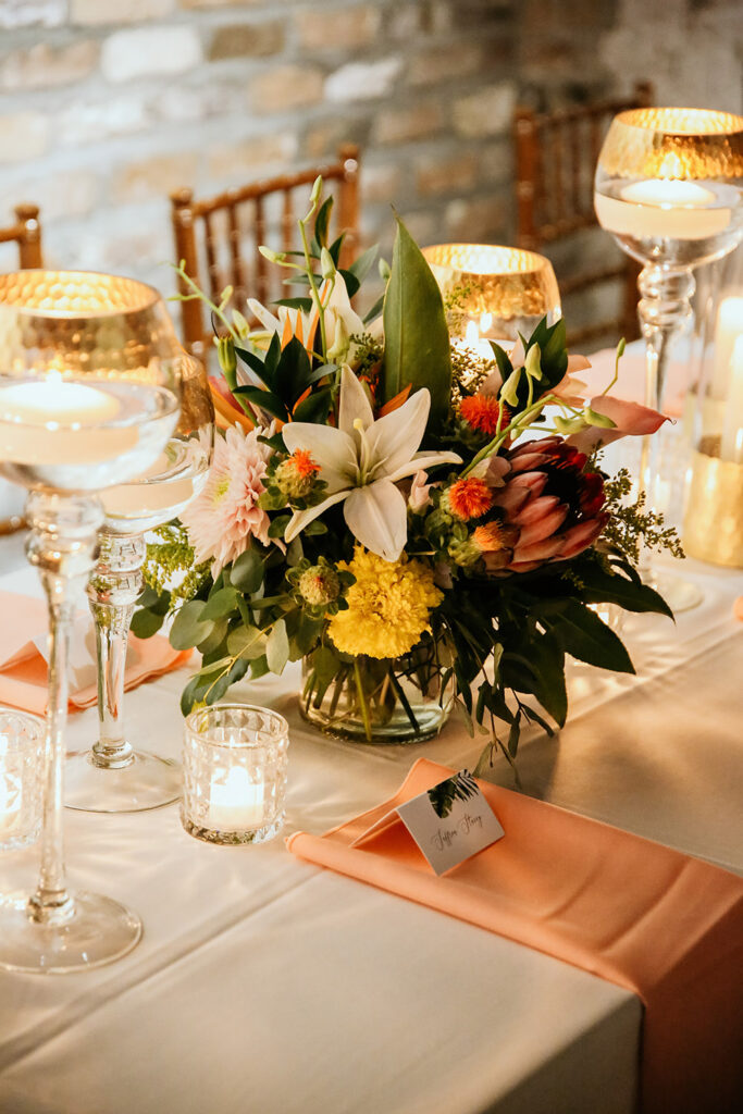 Elegant tropical wedding centerpiece with lilies, protea, marigolds, and greenery surrounded by glowing candles on a reception table with peach napkins at a New Orleans wedding celebration.
