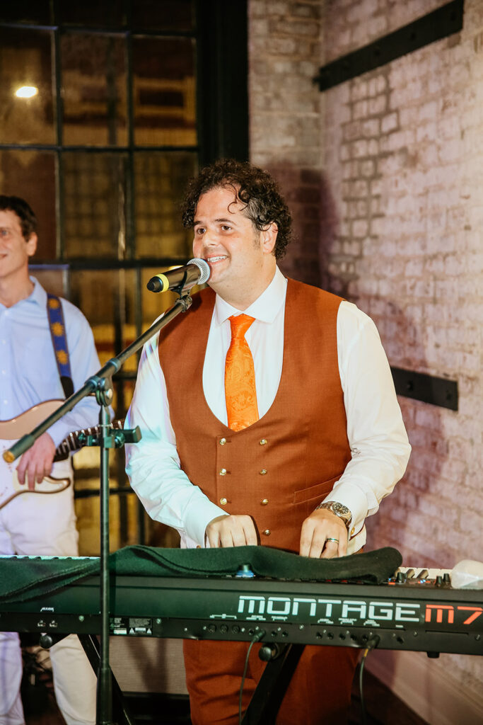 Groom smiles while singing and playing keyboard during the wedding reception, wearing a rust-colored vest and orange tie, with a brick wall and bandmate in the background.