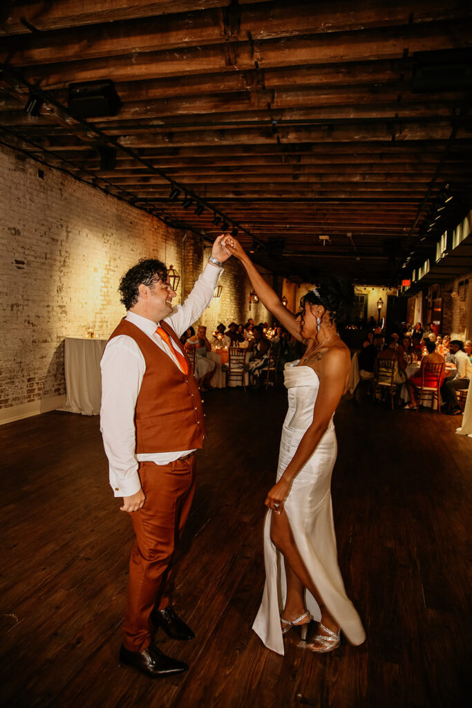 Bride and groom share their first dance at their New Orleans wedding reception, smiling as he twirls her on the wooden dance floor in a brick industrial-style venue.