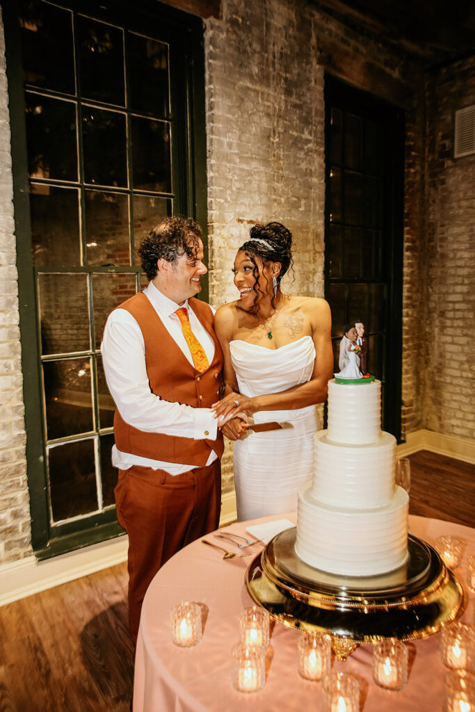 Bride and groom smiling while cutting their white tiered wedding cake at their New Orleans reception, surrounded by candlelight and exposed brick décor.