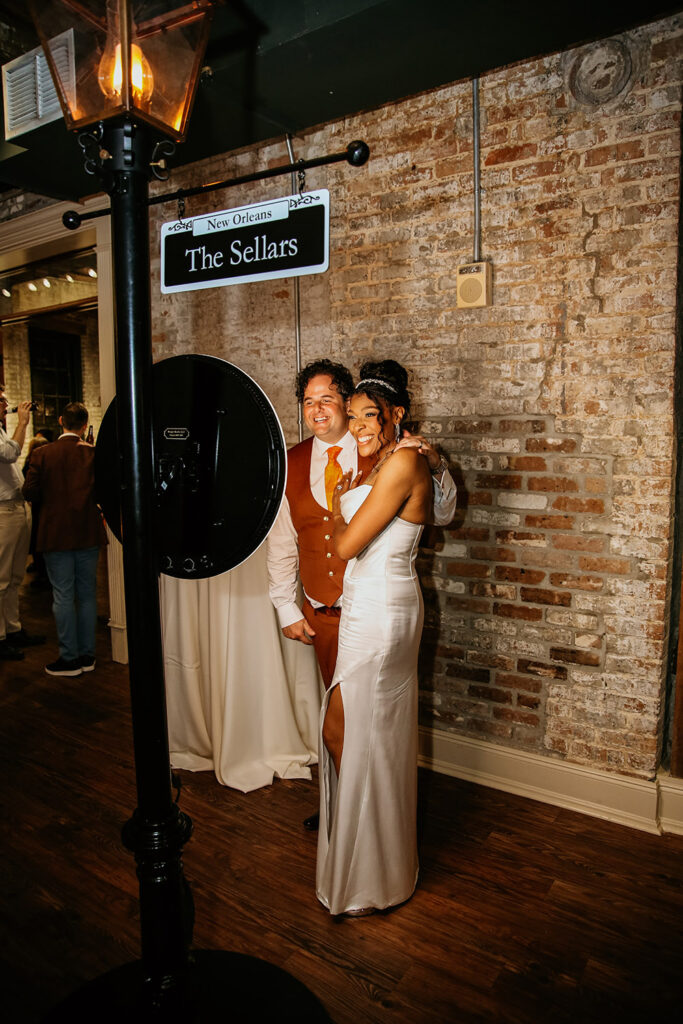 Bride and groom smile together while posing at a photo booth during their New Orleans wedding reception, standing beneath a custom “The Sellars” street-style sign against a rustic brick wall.