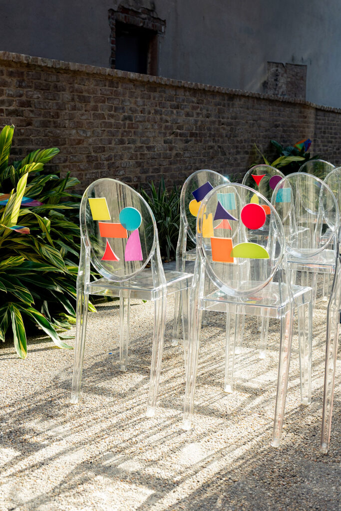 Clear acrylic ceremony chairs decorated with colorful geometric accents, arranged outdoors along a gravel aisle beside greenery and brick walls.