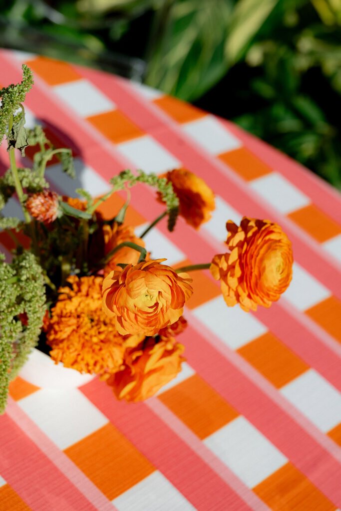 Close-up of orange ranunculus and marigold blooms arranged on a pink and orange checkered tablecloth outdoors.
