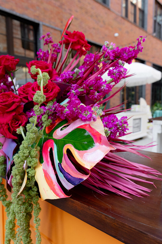 Close-up of a vibrant floral arrangement with red roses, magenta blooms, painted monstera leaves, and textured greenery displayed on an outdoor bar counter.