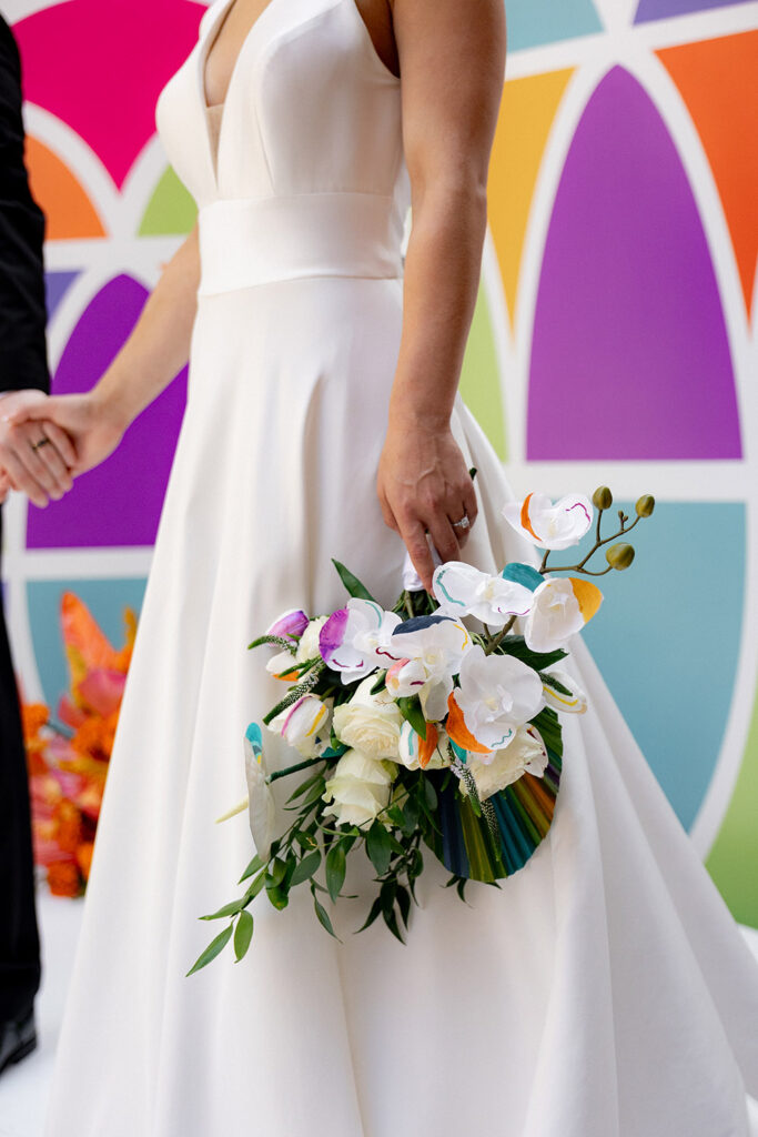 Bride holding a modern white-and-color-accented orchid bouquet while wearing a sleek V-neck wedding gown, standing in front of a colorful geometric ceremony backdrop.