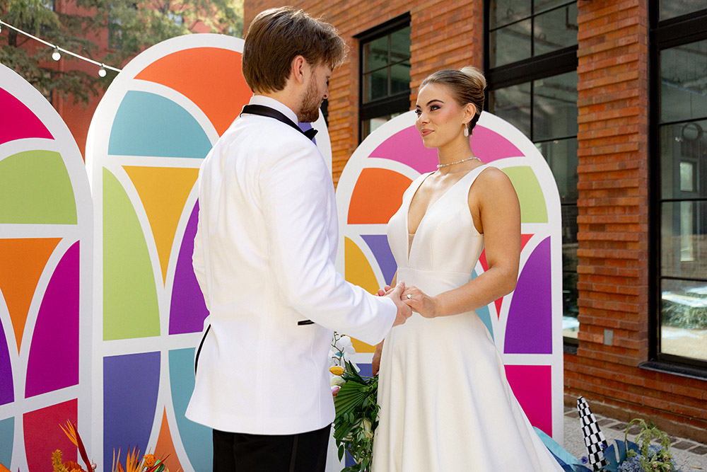 Bride and groom hold hands during their outdoor ceremony in front of a colorful geometric backdrop set between brick buildings.
