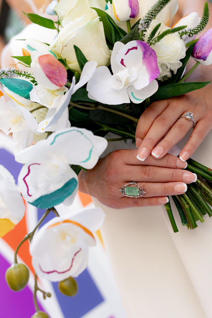 Close-up of a bride’s hands holding a colorful orchid bouquet, showcasing her engagement ring and manicured nails against a vibrant backdrop.