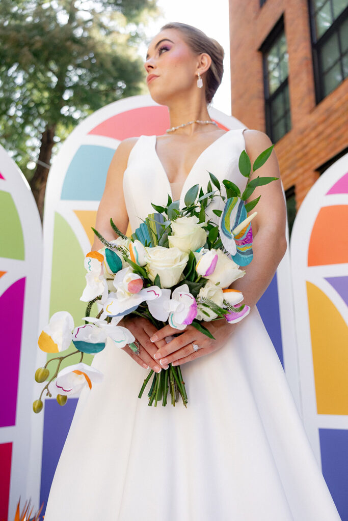 Bride in a sleeveless white gown holds a colorful orchid and rose bouquet in front of a bold geometric ceremony backdrop outdoors.