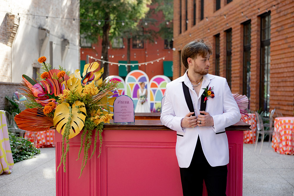 Groom in a white tuxedo jacket stands beside a pink outdoor bar with a bold tropical floral arrangement in a modern courtyard setting.