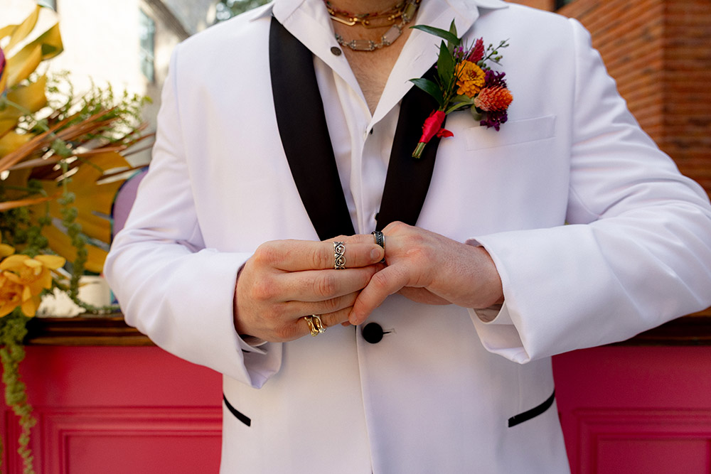 Close-up of groom’s hands buttoning a white tuxedo jacket with black lapels, layered rings, and a colorful boutonniere.