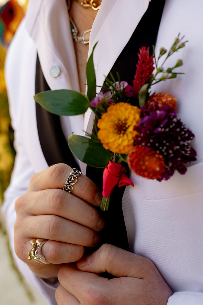 Close-up of groom’s colorful boutonniere pinned to a white tuxedo jacket, with marigold blooms, greenery, and layered statement rings.