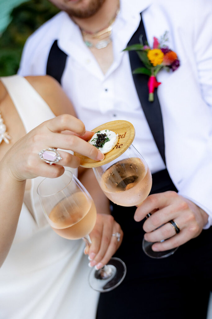 Bride and groom hold champagne flutes while sharing a bite topped with caviar, highlighting wedding rings and modern reception details.