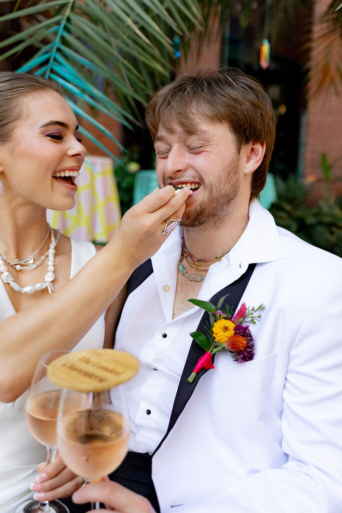 Bride feeds the groom a bite during cocktail hour as they laugh together, holding champagne flutes and wearing colorful boutonnieres.