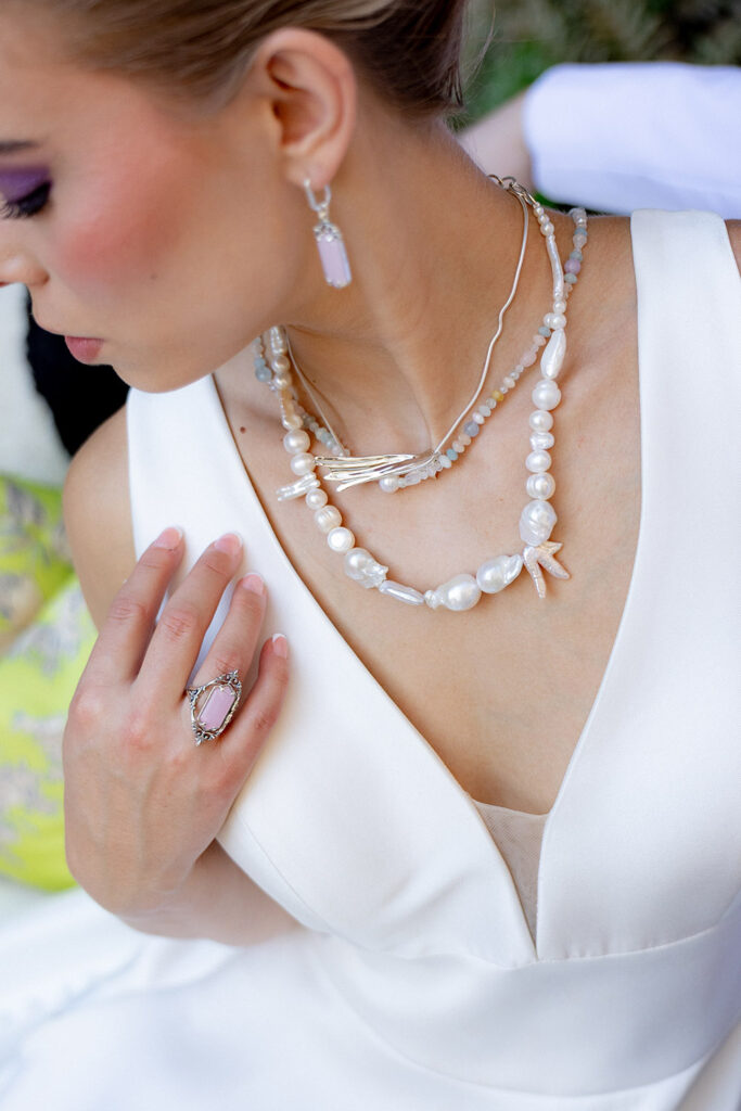Close-up of a bride wearing layered pearl necklaces, a silver necklace, drop earrings, and a gemstone ring with a modern V-neck wedding dress.