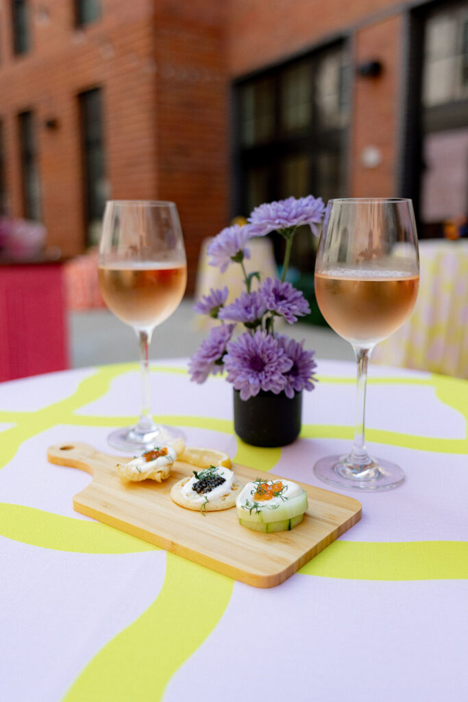 Two glasses of rosé wine on a patterned cocktail table with a small floral arrangement and a wooden board of elegant bite-size appetizers.