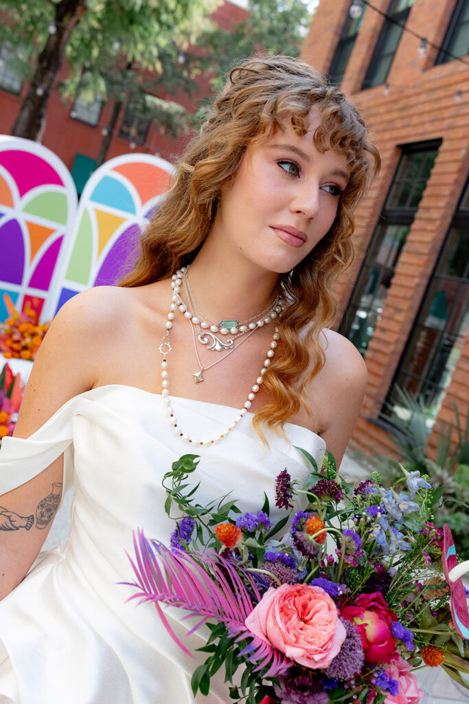 Bride wearing a modern off-the-shoulder wedding gown and layered pearl necklaces holds a vibrant, colorful bouquet in an outdoor courtyard.