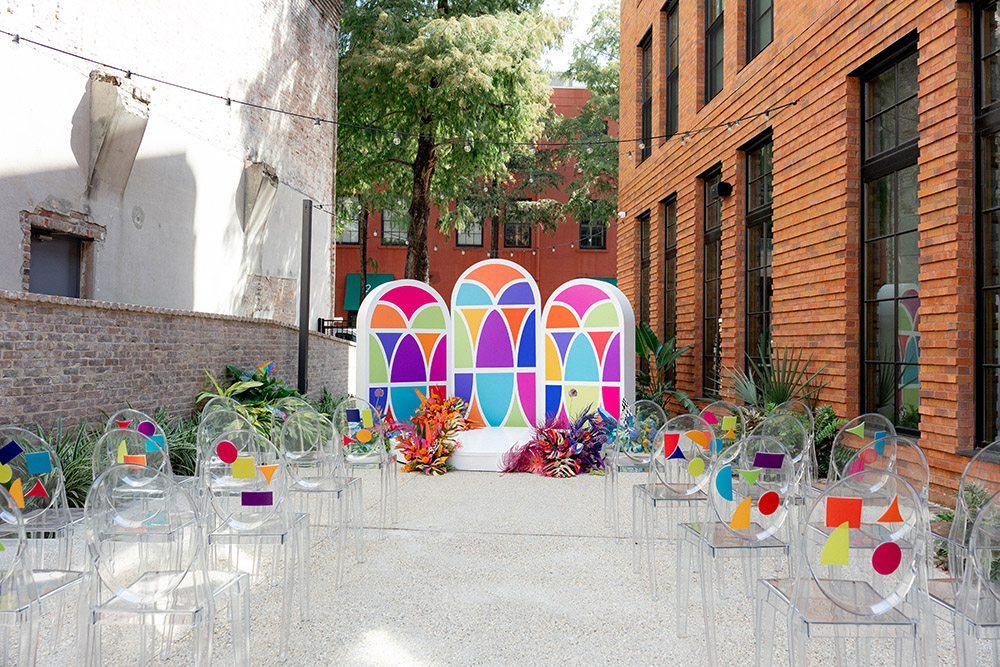 Outdoor wedding ceremony setup with clear acrylic chairs, colorful geometric accents, and a vibrant stained-glass–inspired backdrop between brick buildings.