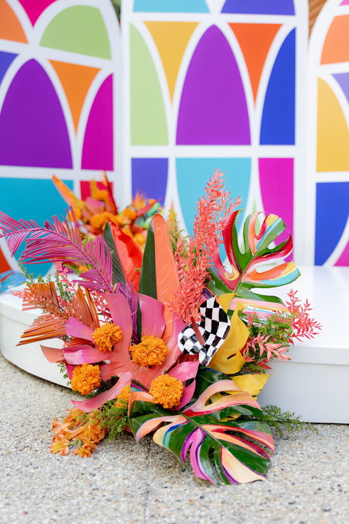 Vibrant ceremony floral installation with painted monstera leaves, tropical blooms, and bold colors arranged at the base of a colorful geometric backdrop.