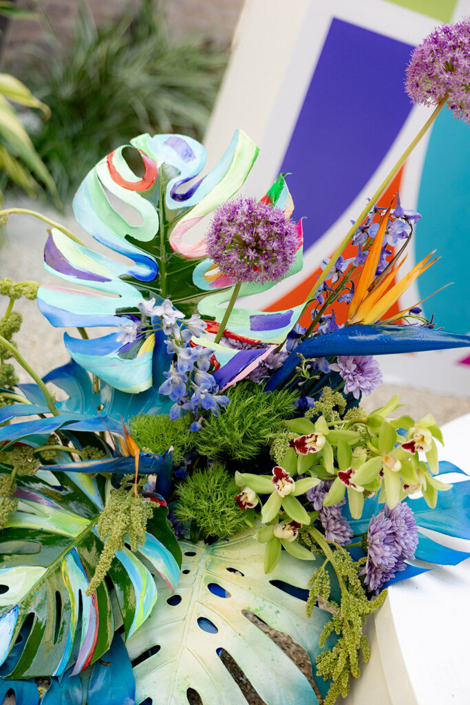 Close-up of a bold, colorful floral arrangement with painted monstera leaves, orchids, allium blooms, and tropical accents in vibrant blues, greens, and purples.