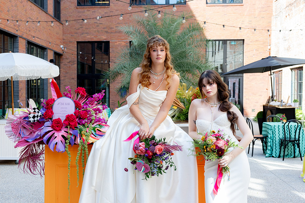 Two brides pose in modern wedding gowns holding vibrant bouquets in an outdoor courtyard with bold florals, palm greenery, and string lights.