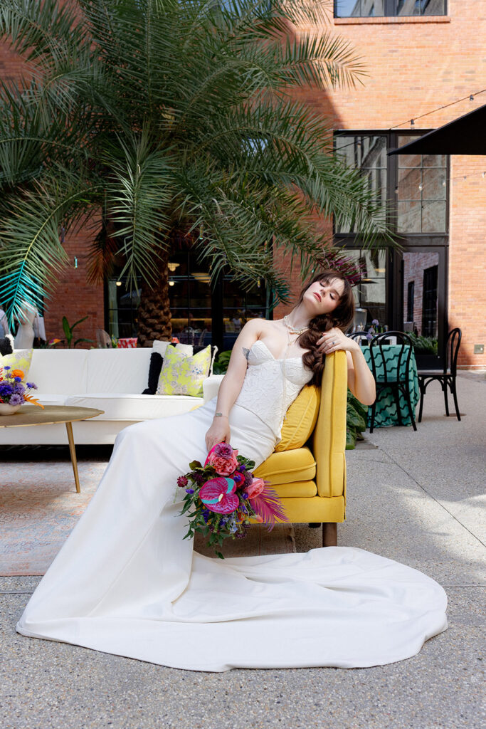 Bride in a fitted wedding gown lounges on a yellow chair, holding a bold, colorful bouquet in a modern outdoor courtyard with palm trees.