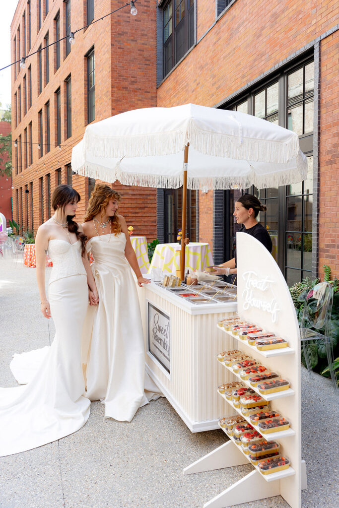 Two brides in white wedding gowns stand together at a dessert cart with individual cheesecake cups under a white umbrella in an outdoor courtyard.