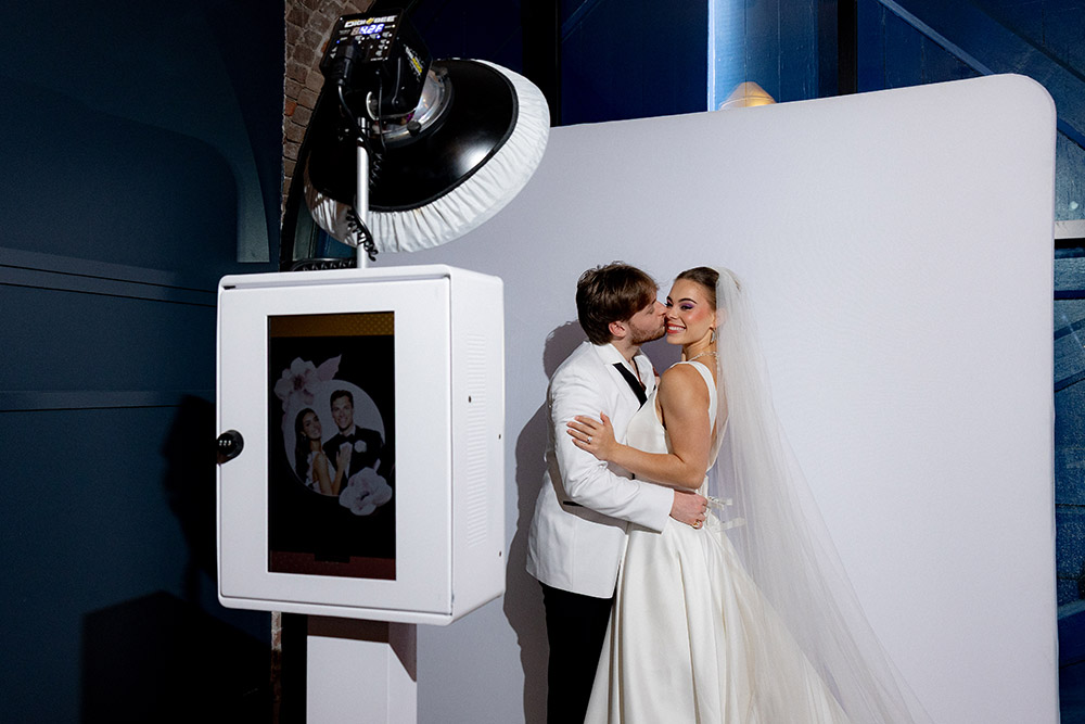 Bride and groom pose and kiss at a modern wedding photo booth with a white backdrop and built-in camera during their reception.